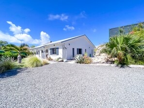 Une maison blanche avec une allée en gravier, entourée de différentes plantes et sous un ciel bleu clair.