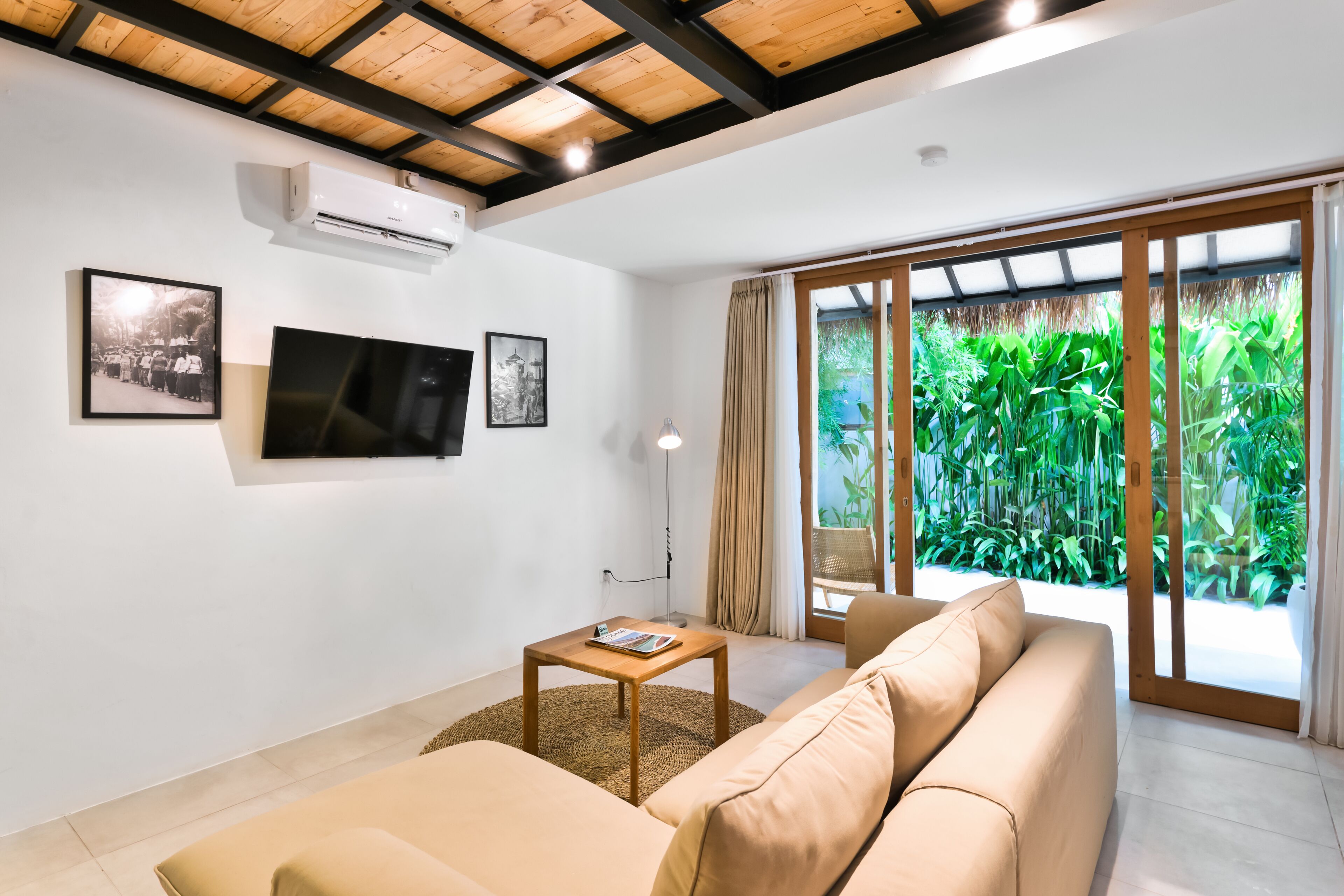 Ein modernes Wohnzimmer mit einem beigefarbenen Sofa, einem Holztisch und einem flachbildfernseher an der Wand. Eine große Glastür öffnet sich zu einem üppigen, grünen Garten.