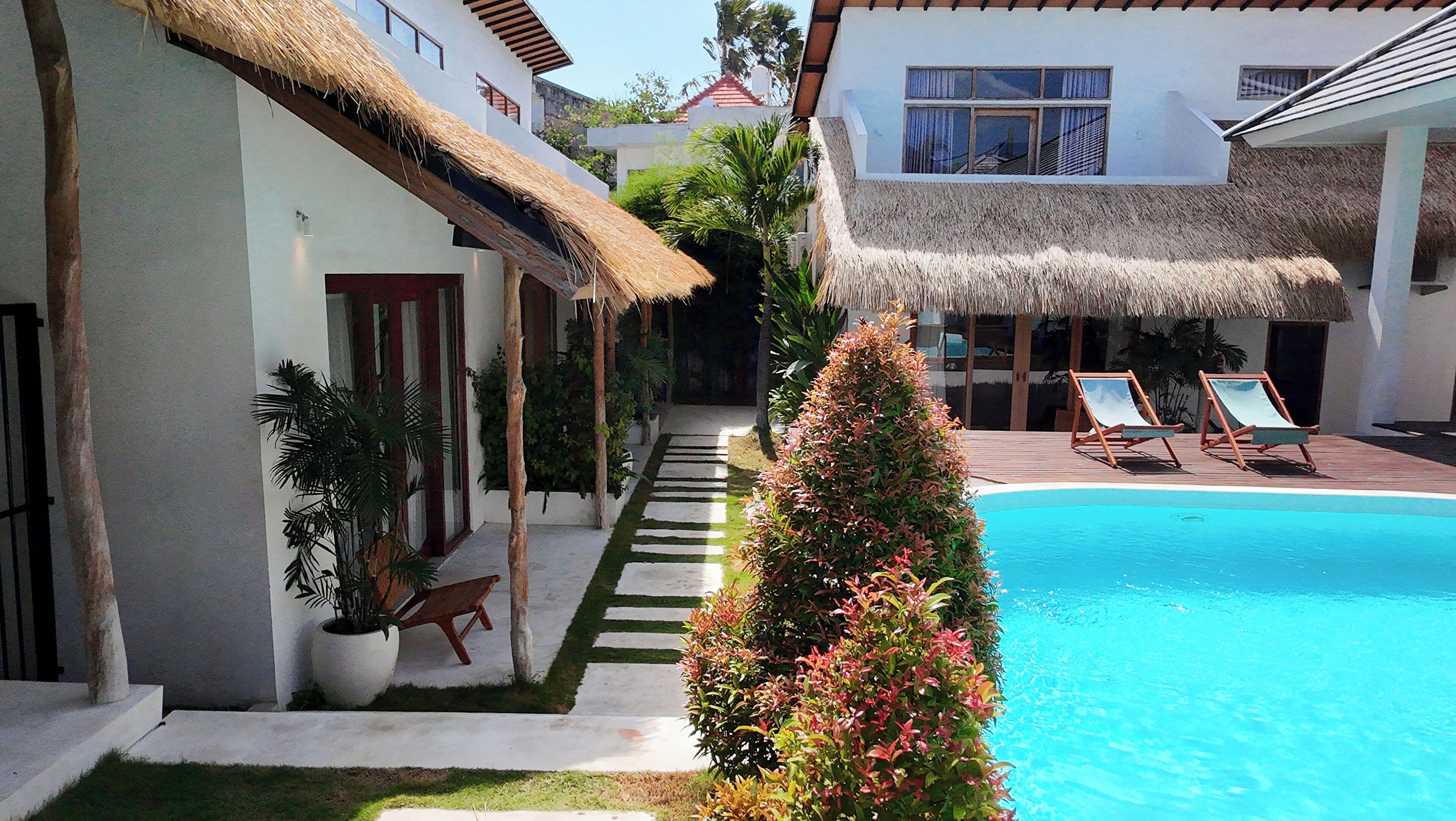 A poolside area with a clear blue pool, lounge chairs, and a thatched-roof building.