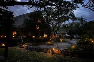 A rustic building with stone walls and a thatched roof, surrounded by trees and a small pond with a stone wall.