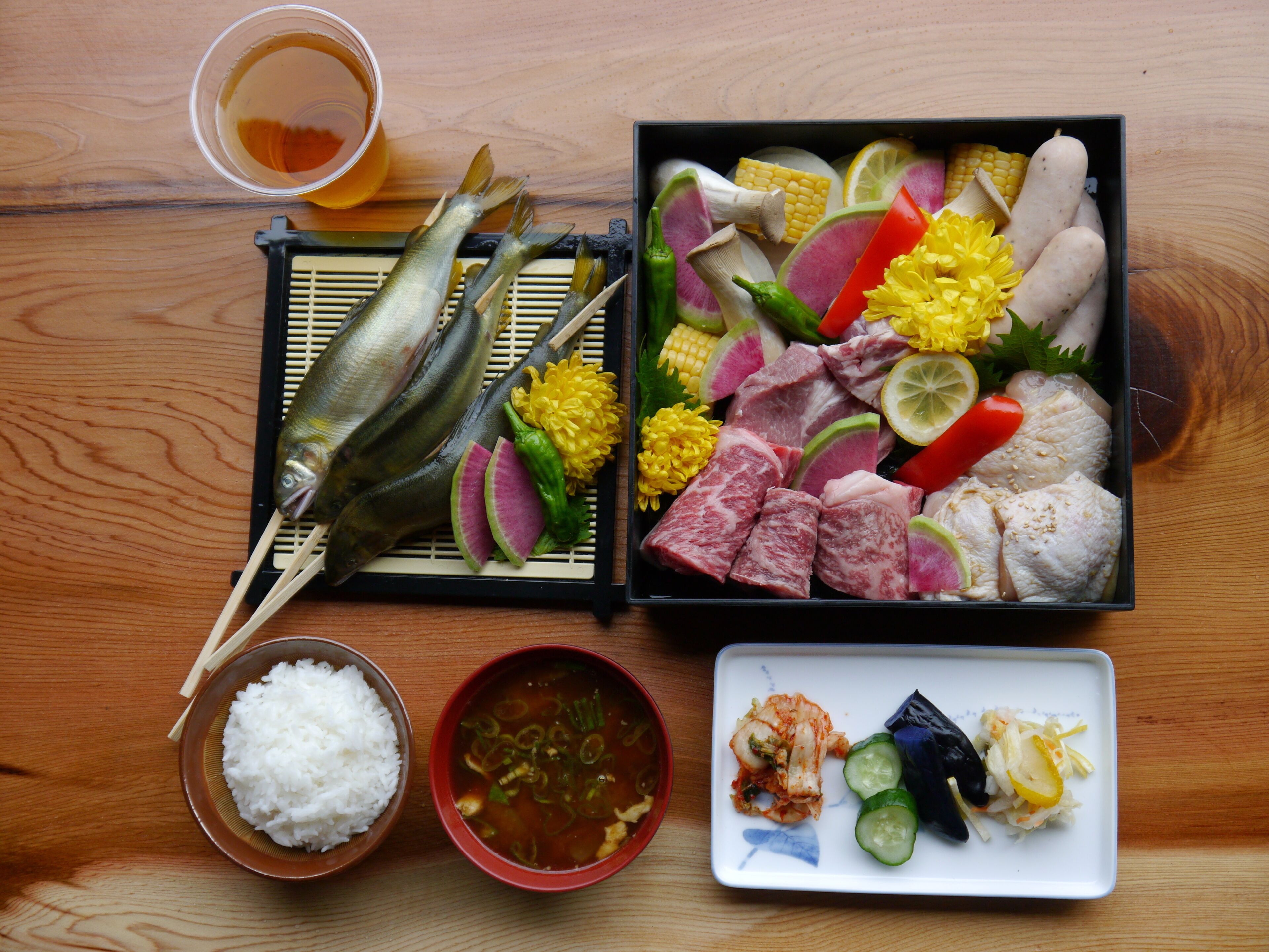 Une table en bois sur laquelle est servi un repas composé de poisson, de viande, de légumes et de riz, accompagné d’une tasse de thé.