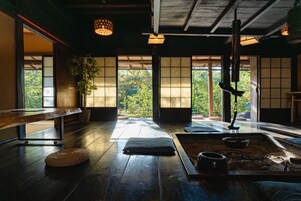 A traditional Japanese room with tatami flooring, a low table, a hanging kettle, and a view of greenery through large windows.