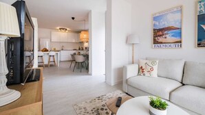 A modern living room with a sofa, a coffee table, and a TV. There is a kitchen in the background with white cabinets and a dining area. - Seascape Apartment Unit 2 by Portugal Portfolio (Estoril)