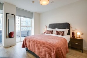 A modern bedroom with a large bed, a nightstand with a lamp, and a view of the city through large windows.