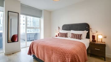 A modern bedroom with a large bed, a nightstand with a lamp, and a view of the city through large windows.