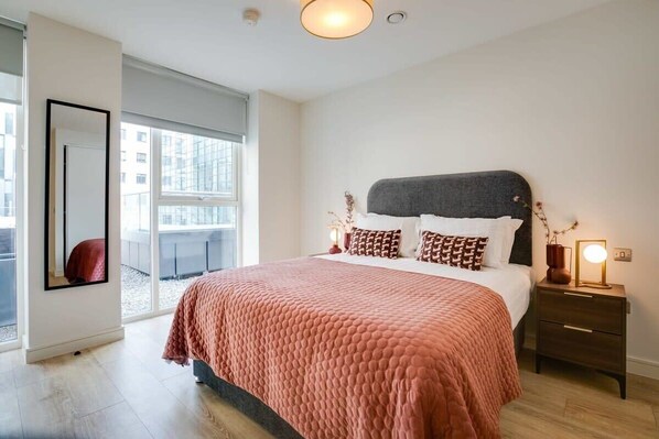 A modern bedroom with a large bed, a nightstand with a lamp, and a view of the city through large windows.