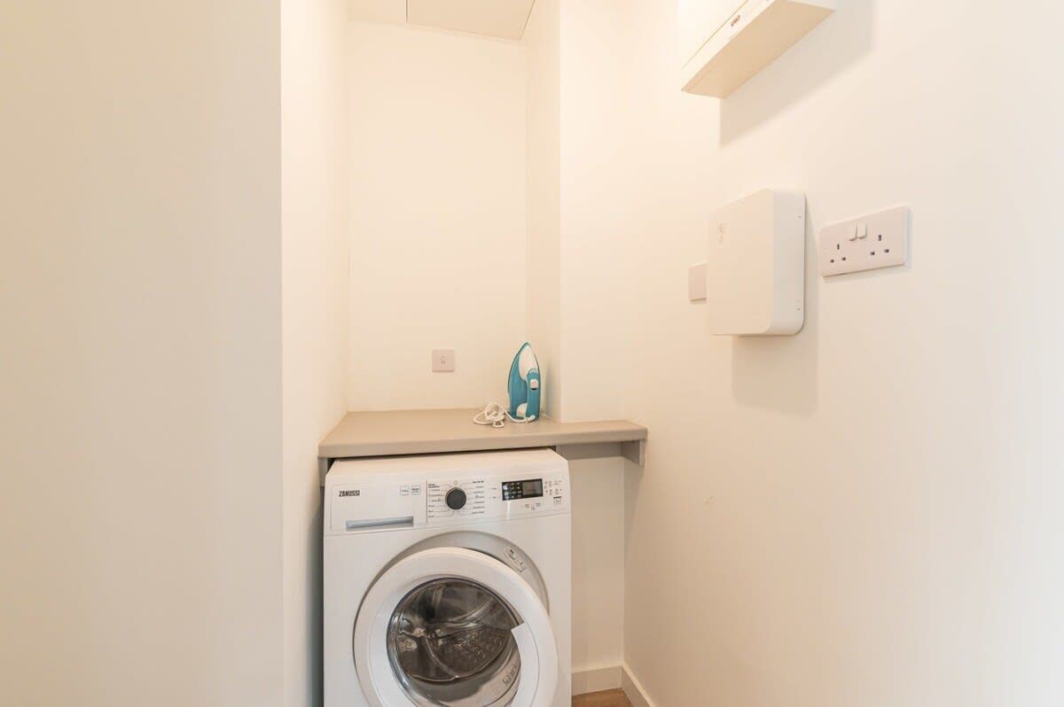 Une petite buanderie équipée d’un lave-linge à chargement frontal, d’une petite étagère sur laquelle se trouve un appareil électroménager bleu, et d’une prise électrique murale.