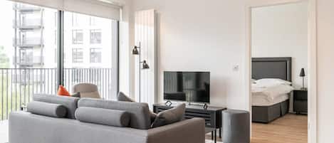 A modern living room with a grey sofa, a flat-screen TV, and a bed with a white duvet.