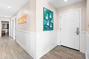 A hallway with a white door, a painting of sailboats, and a kitchen area in the background.