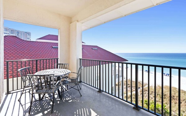 A balcony with a table and chairs, overlooking a beach and buildings with red roofs.