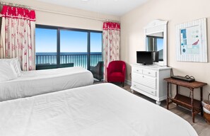 A hotel room with two beds, a television on a dresser, a red chair, and a view of the ocean.