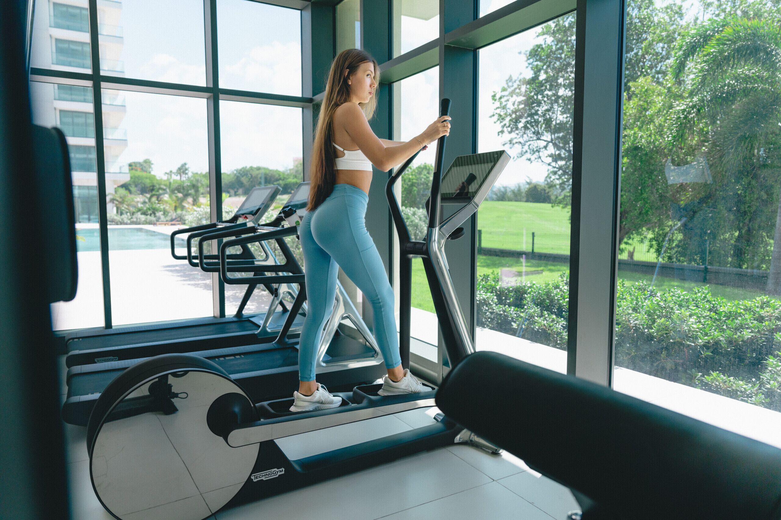 A person using a stationary bike in a gym with large windows and exercise equipment in the background.