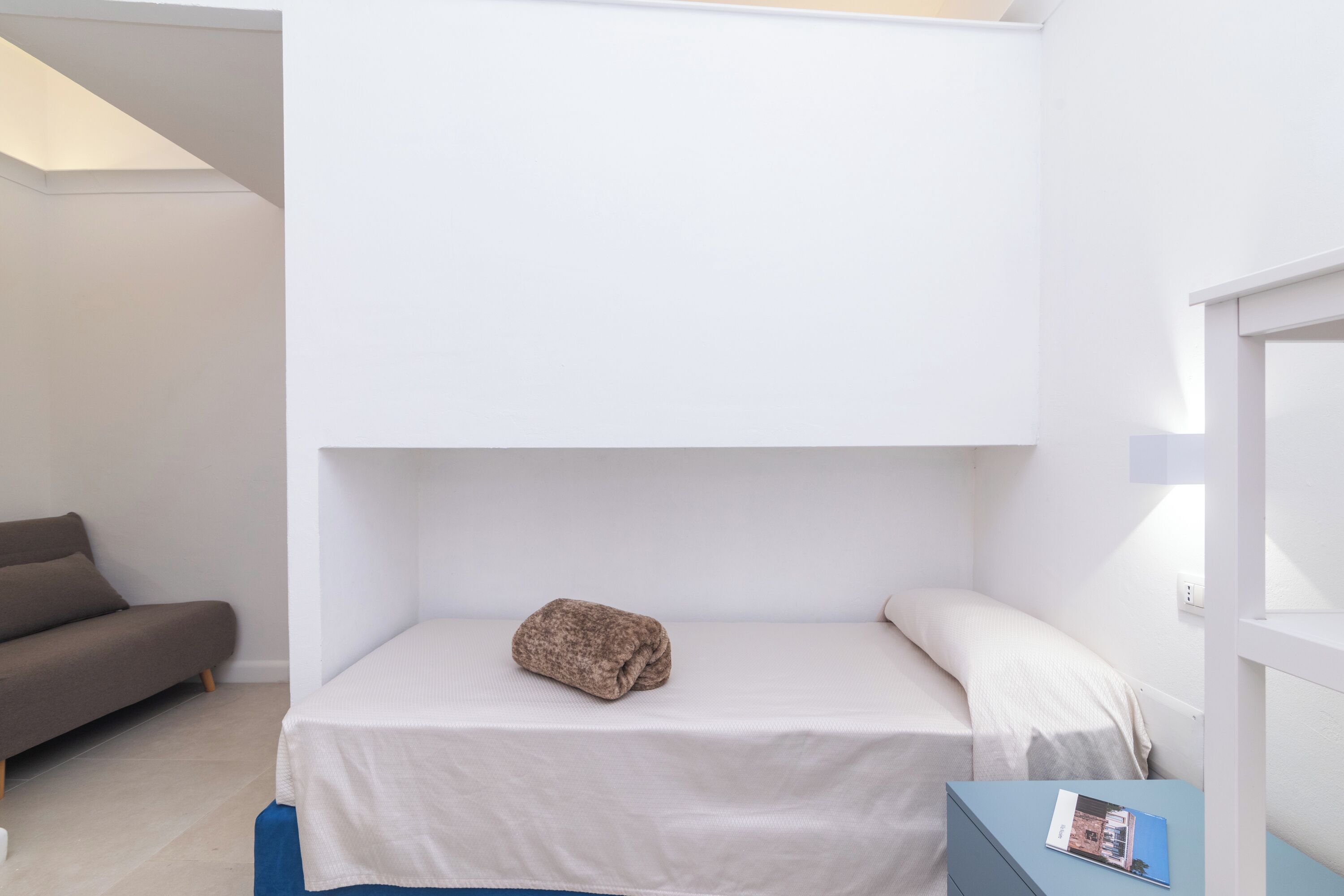A bunk bed with a folded blanket on the lower bunk, a small table with a magazine, and a sofa in the adjacent room.