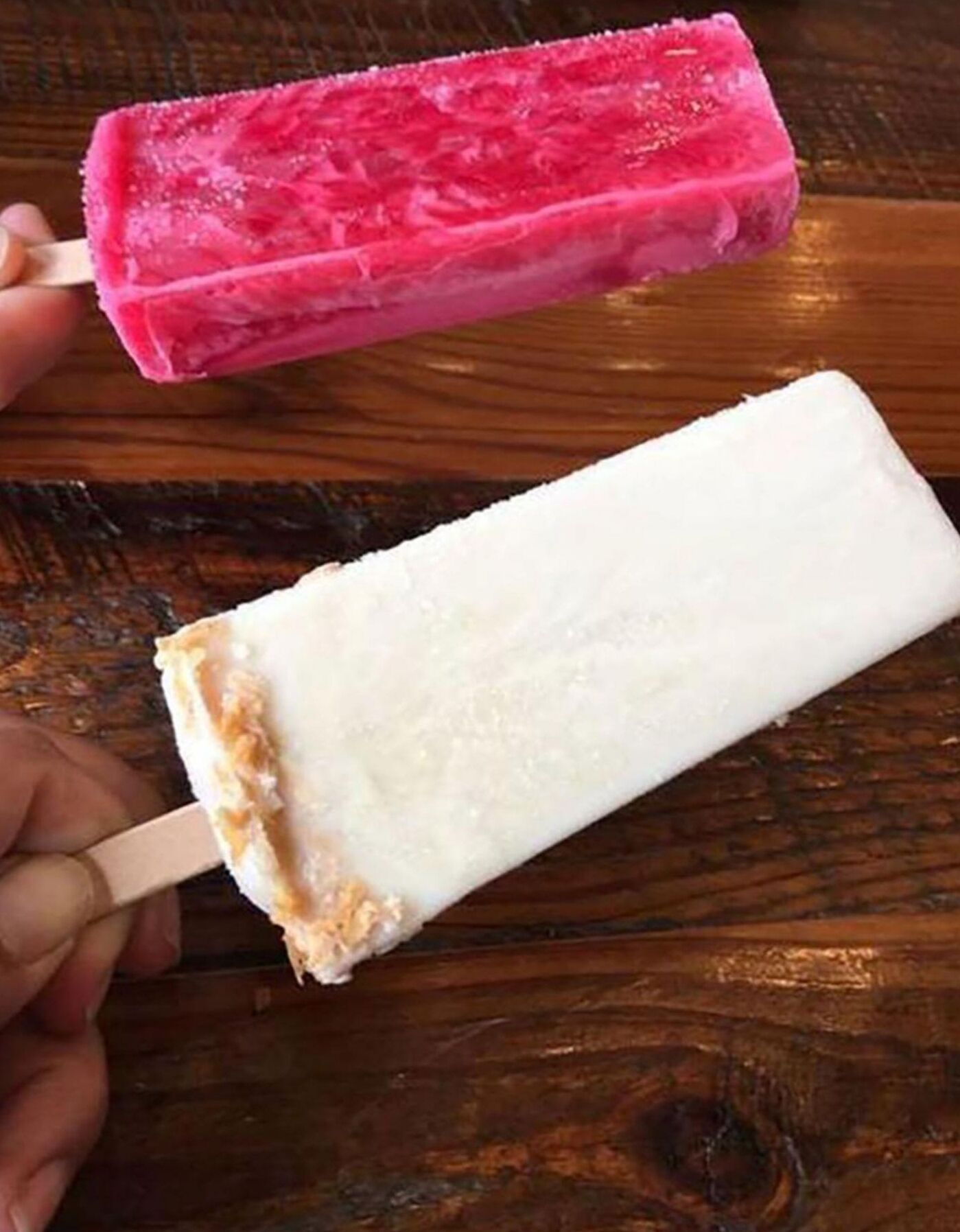 Uma mão segura dois sorvetes em palito, um rosa e um branco.