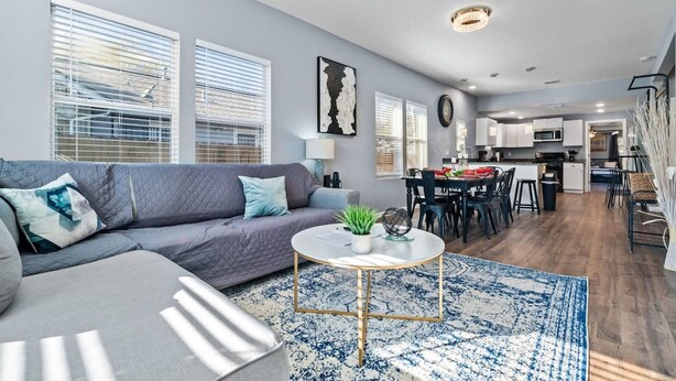 Een moderne woonkamer met een grijze bank, een ronde salontafel en een blauw vloerkleed. Er is een eethoek met een tafel en stoelen, en een keuken op de achtergrond.