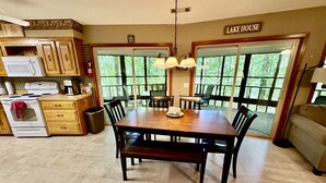 Un coin repas avec une table et des chaises en bois, une cuisine avec des meubles et des appareils électroménagers en bois, et de grandes fenêtres donnant sur la forêt.