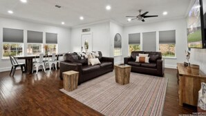 A spacious living room with a dark brown leather sofa, a wooden coffee table, and a striped rug. There are two windows with blinds, a ceiling fan, and a flat-screen TV mounted on the wall. - Experience Serenity in a 4BR 3BA Downtown Home (San Antonio)