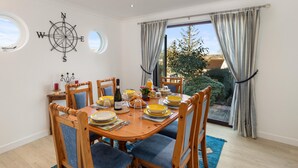 A dining room with a wooden table set for six, blue upholstered chairs, a compass wall decoration, and a view of the outdoors through large windows. - Four Oaks - Sea and Harbour Views (Saundersfoot)