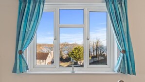 A window with teal curtains, a white frame, and a view of a landscape with trees and buildings. - Four Oaks - Sea and Harbour Views (Saundersfoot)