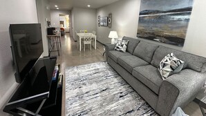 A modern living room with a flat-screen TV, grey sofa, and a painting of a landscape. - Brand New Rest and Relaxation Apartments (Clovis)