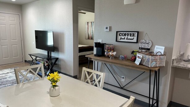 A dining area with a white table and chairs, a vase with yellow flowers, a coffee station with a sign, and a television in the background. - Brand New Rest and Relaxation Apartments (Clovis)