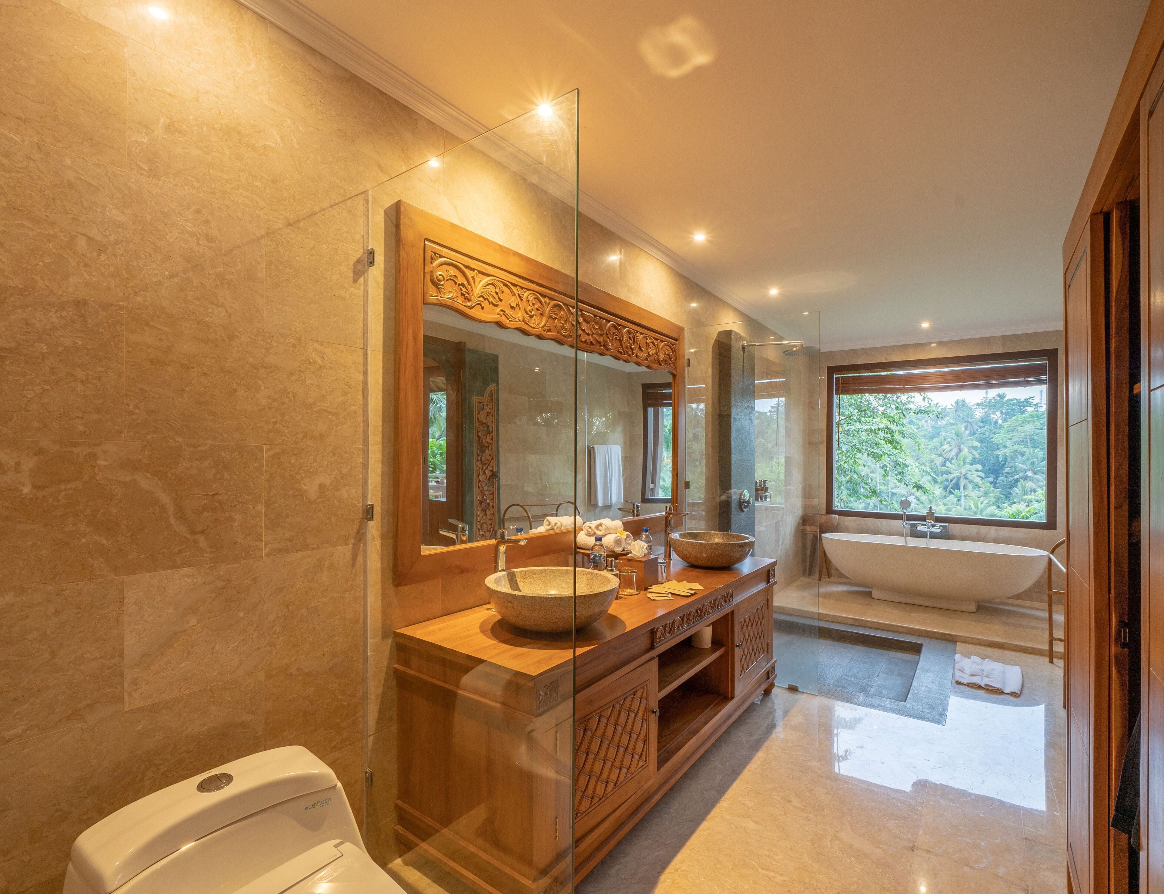 A modern bathroom with a large mirror, a wooden vanity, a freestanding bathtub, and a view of greenery outside.