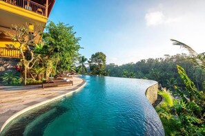 A curved infinity pool with a wooden deck and lounge chairs, surrounded by lush greenery and a building with balconies. - GK Bali Resort  (Sebatu)