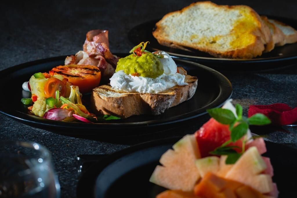 A plate with grilled vegetables, a slice of toast topped with guacamole and cream, and a piece of bread.