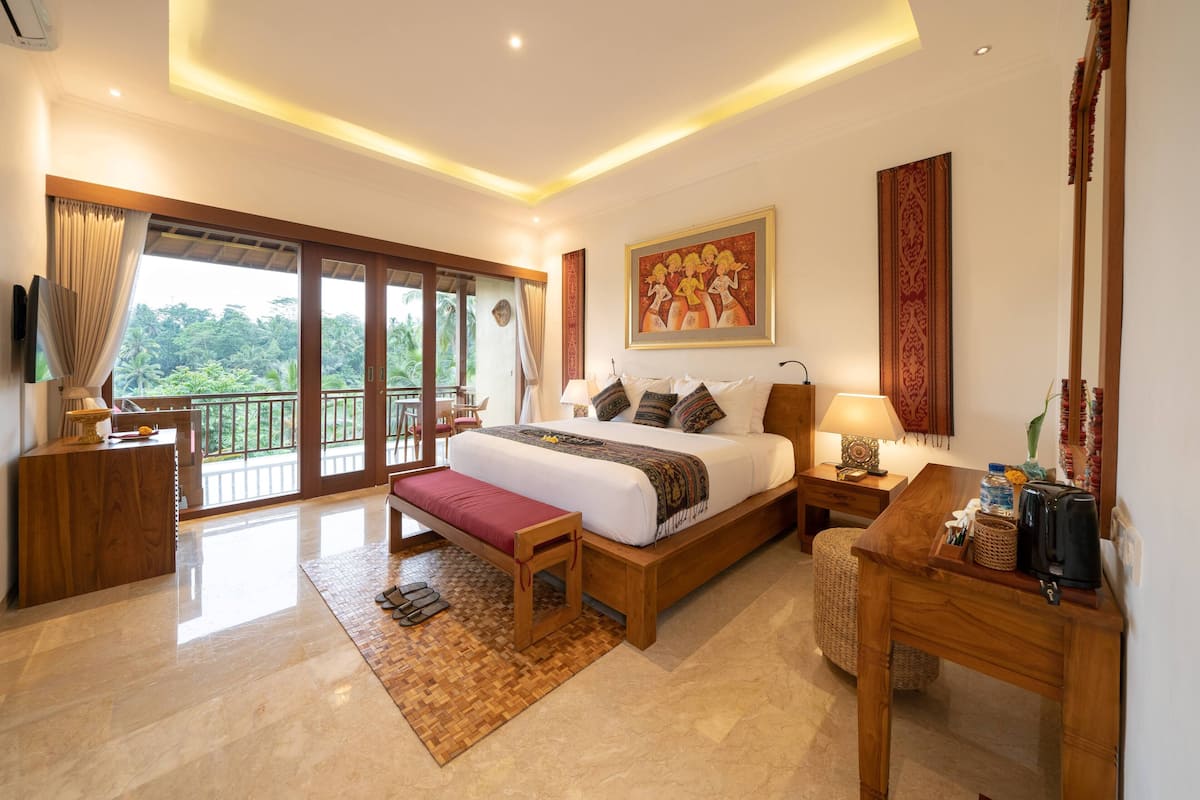 A spacious bedroom with a large bed, a wooden bench, a bedside table with a lamp, and a view of lush greenery through the balcony doors.
