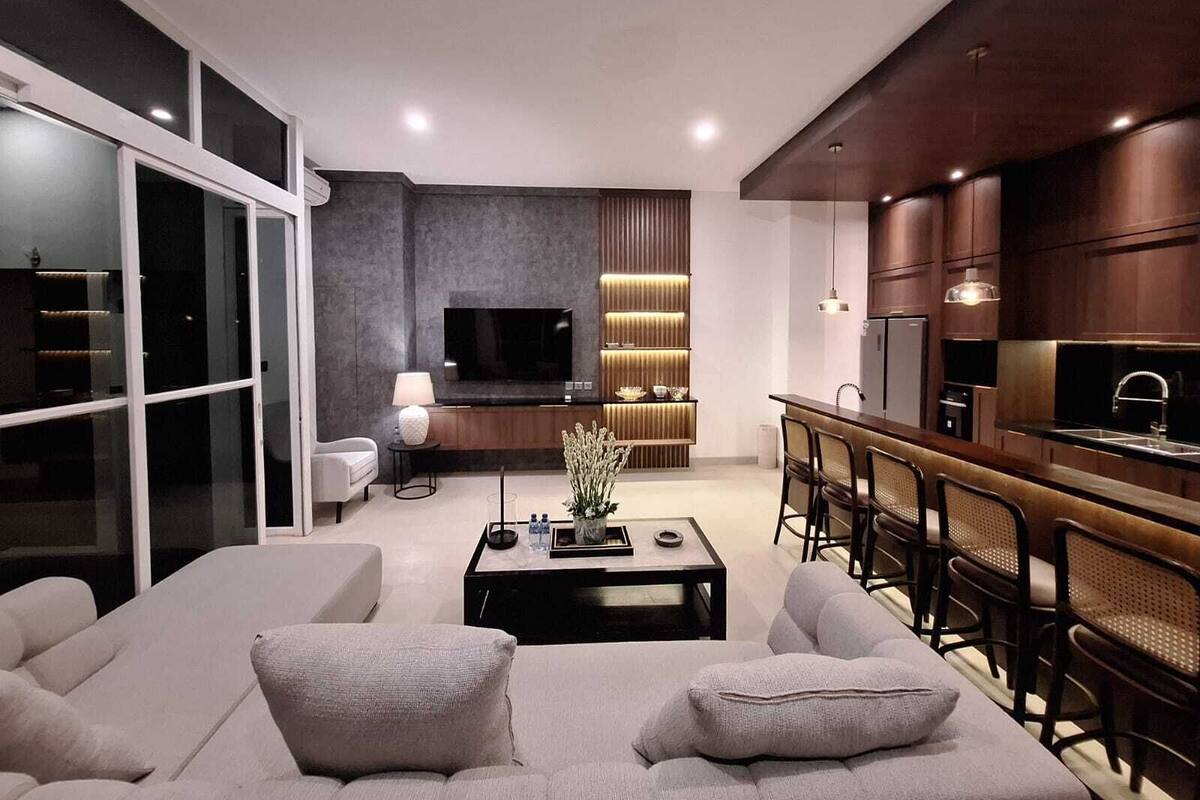 A modern living room with a sofa, coffee table, and a TV. There is a kitchen area with a bar and seating.