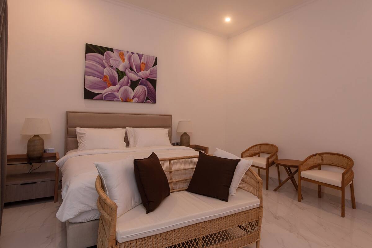 A bedroom with a bed, two nightstands, a wicker sofa, two chairs, and a painting on the wall.