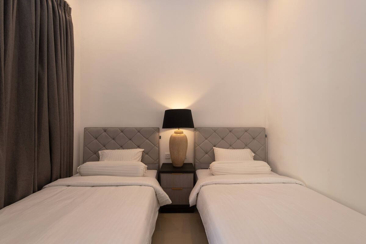 A hotel room with two beds, a nightstand with a lamp, and a large window with curtains.