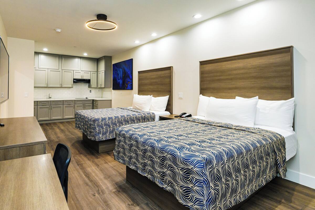 A hotel room with two beds, a desk, a chair, a small table, a kitchen area, and artwork on the walls.
