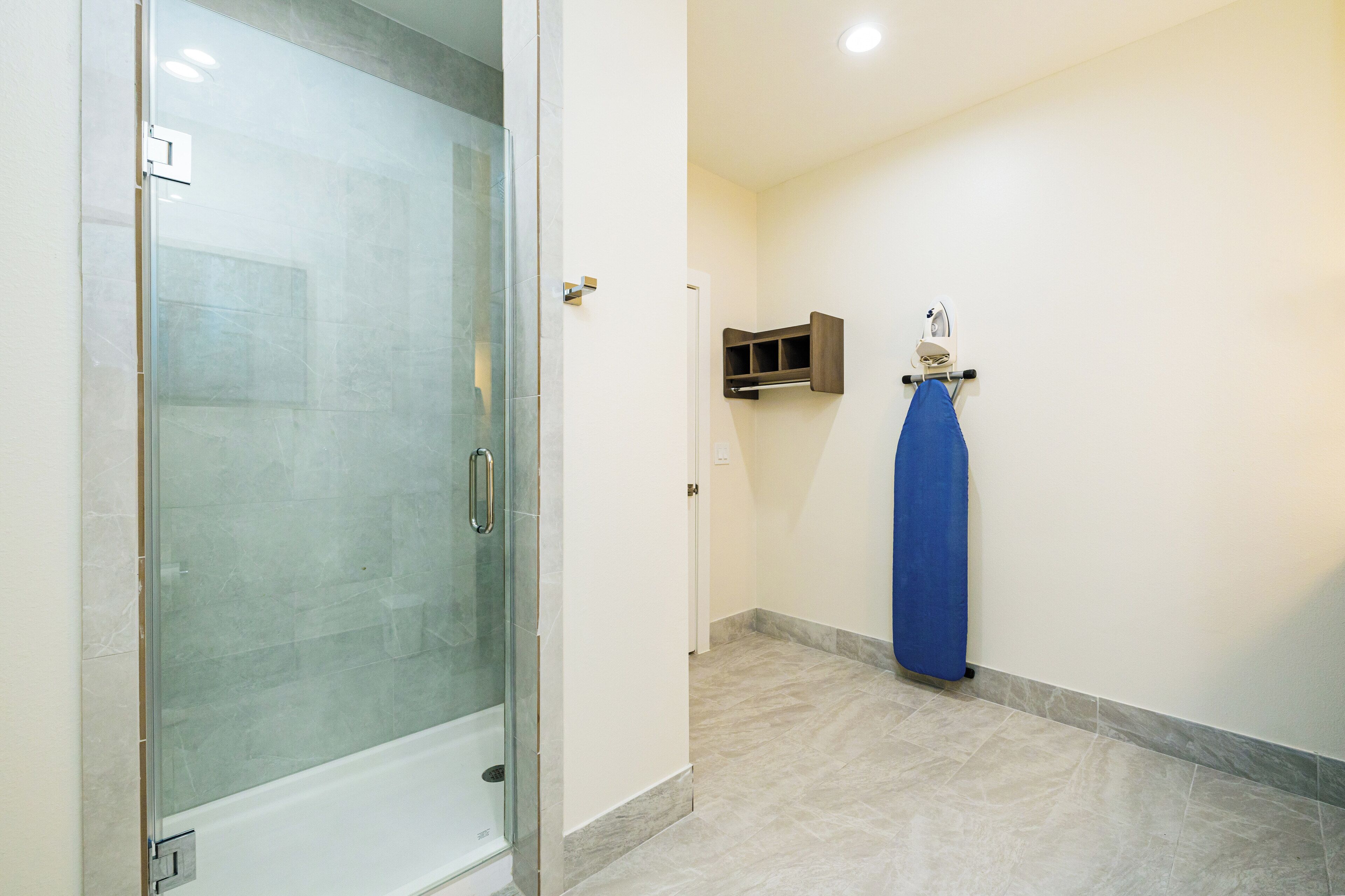 A bathroom with a glass-enclosed shower, a blue towel hanging on a rack, and a small shelf with an iron.