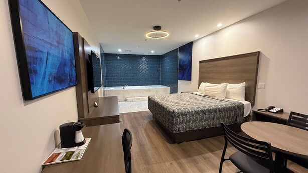 A modern hotel room with a large bed, a desk with a chair, a small table, a sofa, a TV, and a bathroom with a bathtub and shower. -  Palace Inn Blue Edgebrook (Houston)