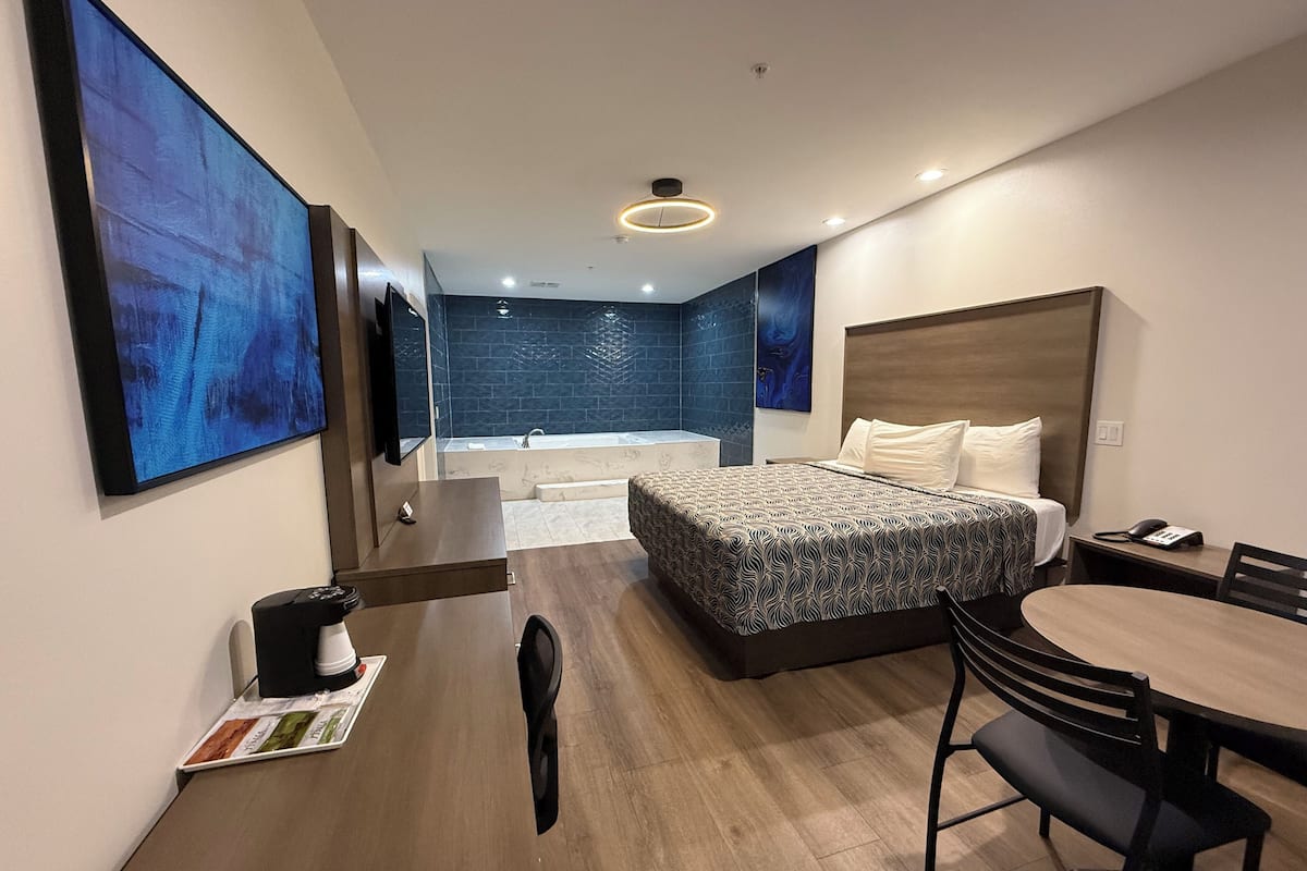A modern hotel room with a large bed, a desk with a chair, a small table, a sofa, a TV, and a bathroom with a bathtub and shower.