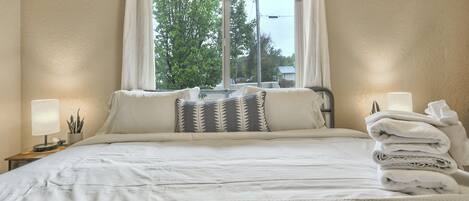 Een net opgemaakt bed met witte lakens, een bedlampje en een stapel handdoeken aan de rechterkant.