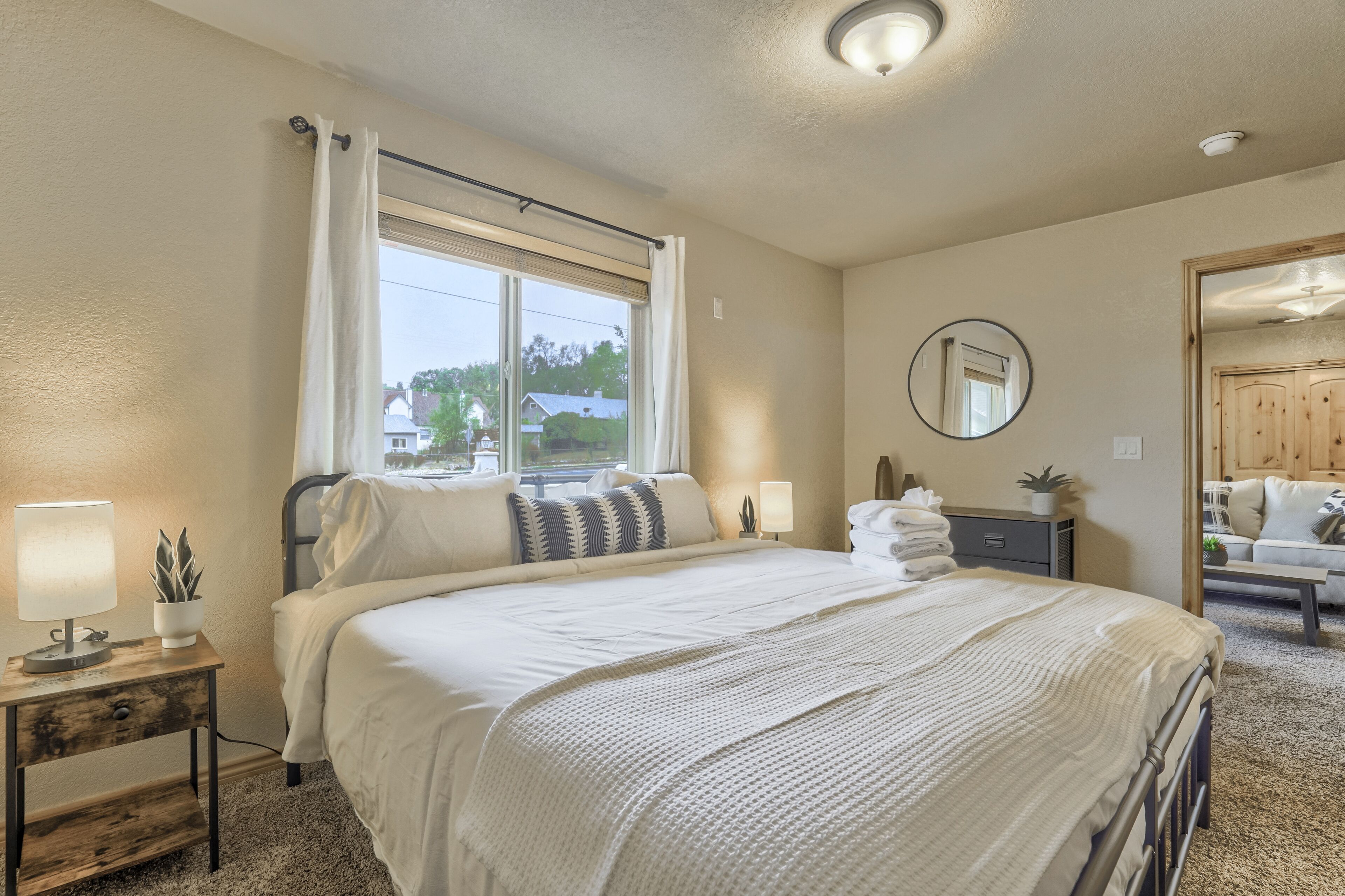A bedroom with a large bed, a bedside table with a lamp, a window with curtains, a mirror on the wall, and a view of the outside.