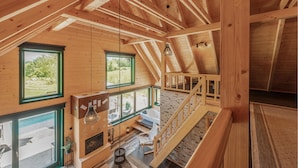 A wooden cabin with a loft bedroom, a fireplace, and large windows offering views of the outdoors.