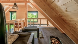 A spacious loft bedroom with a high ceiling, wooden beams, and large windows offering a view of greenery outside.
