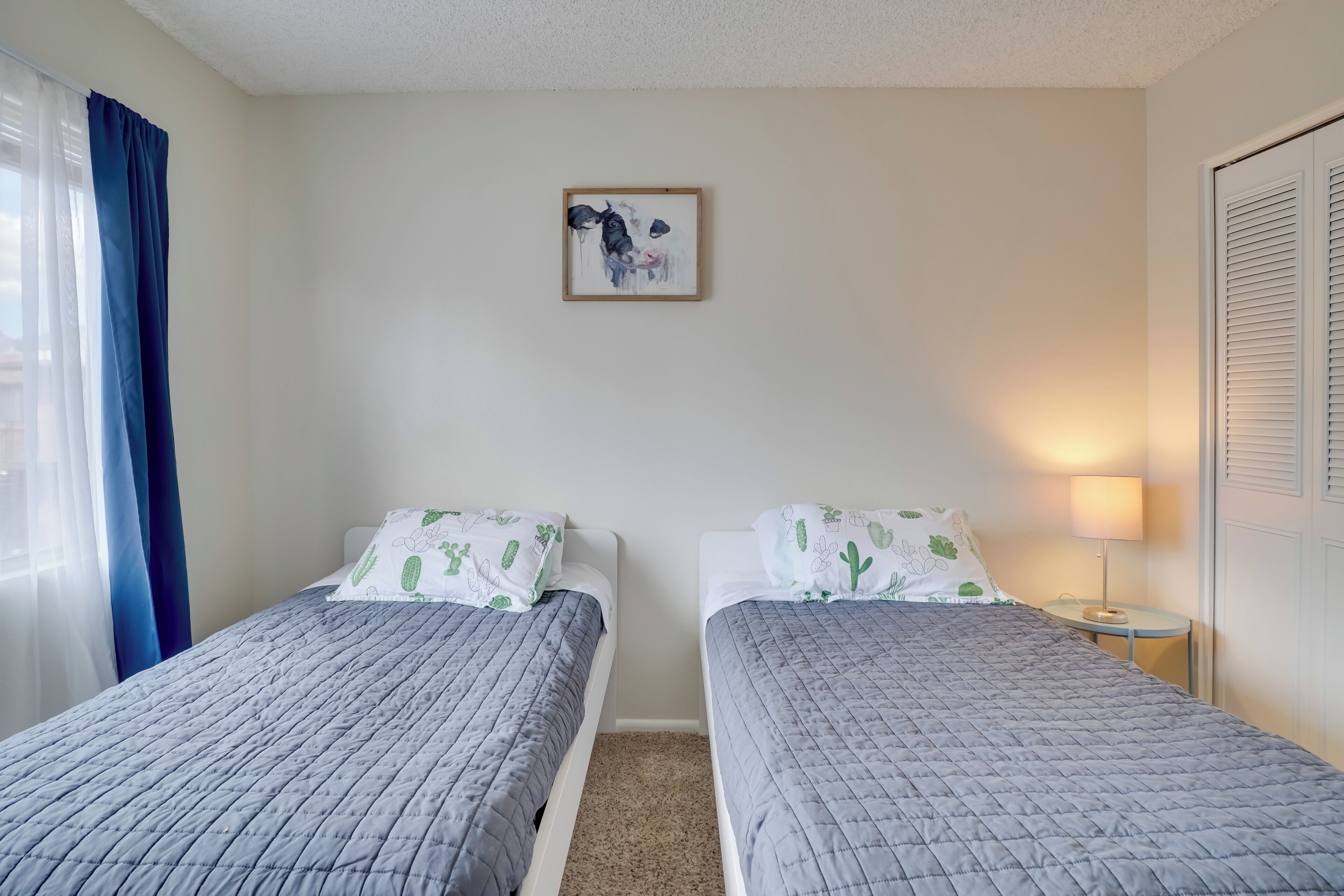 Ein Zimmer mit zwei Betten, einem gerahmten Kunstwerk an der Wand, einer Lampe und einem Fenster mit Vorhängen.