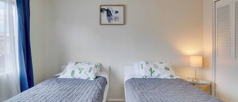 Ein Zimmer mit zwei Betten, einem gerahmten Kunstwerk an der Wand, einer Lampe und einem Fenster mit Vorhängen.