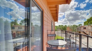 En balkon med glasdør, et metalbord og stole, med udsigt over en parkeringsplads og tilstødende bygninger.