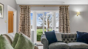 Ein Wohnzimmer mit grauem Sofa, grün-blauen Kissen, blumigen Vorhängen und Blick durch eine Glastür.