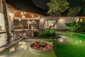 Un espace de restauration aménagé au bord de la piscine, avec une table ronde dressée pour un repas, entouré d’une végétation luxuriante et d’une structure à toit de chaume.