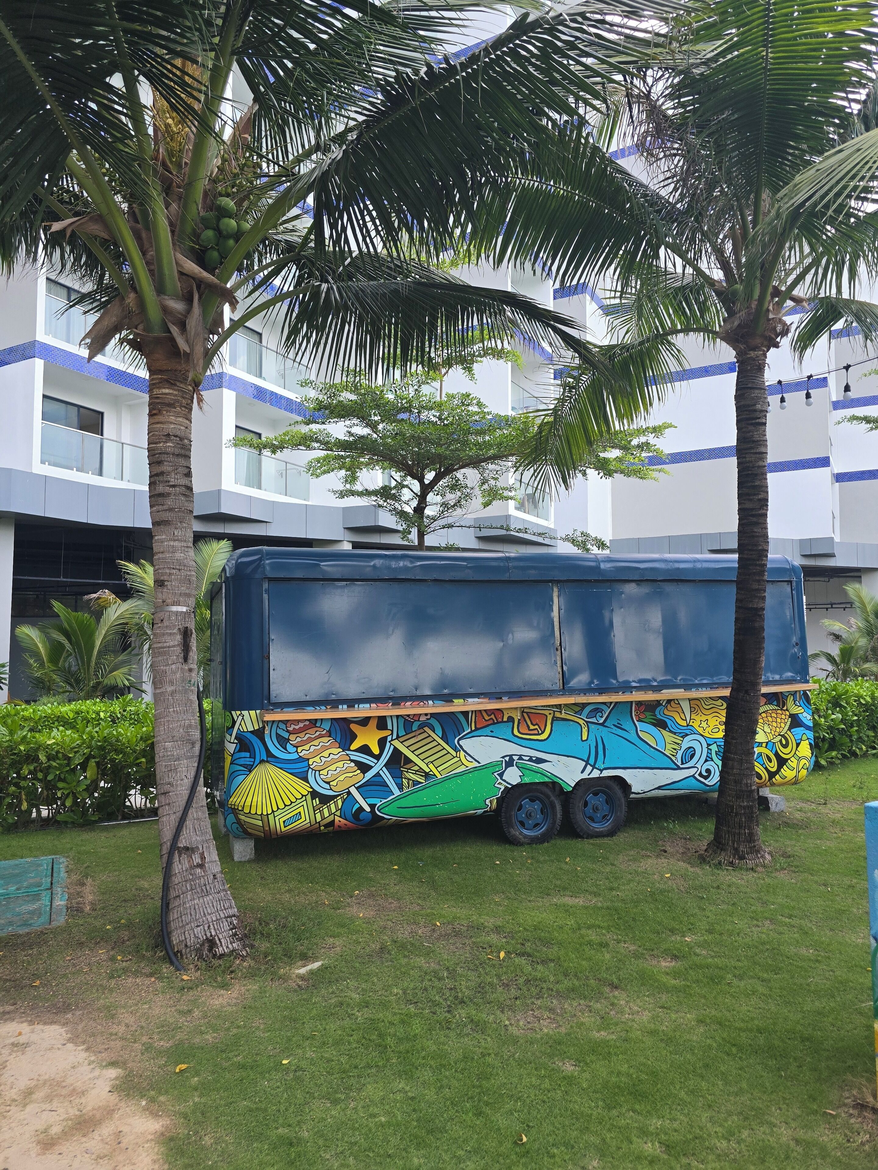 Ein farbenfroher Bus auf einer Rasenfläche, im Hintergrund ein modernes Gebäude und Palmen.