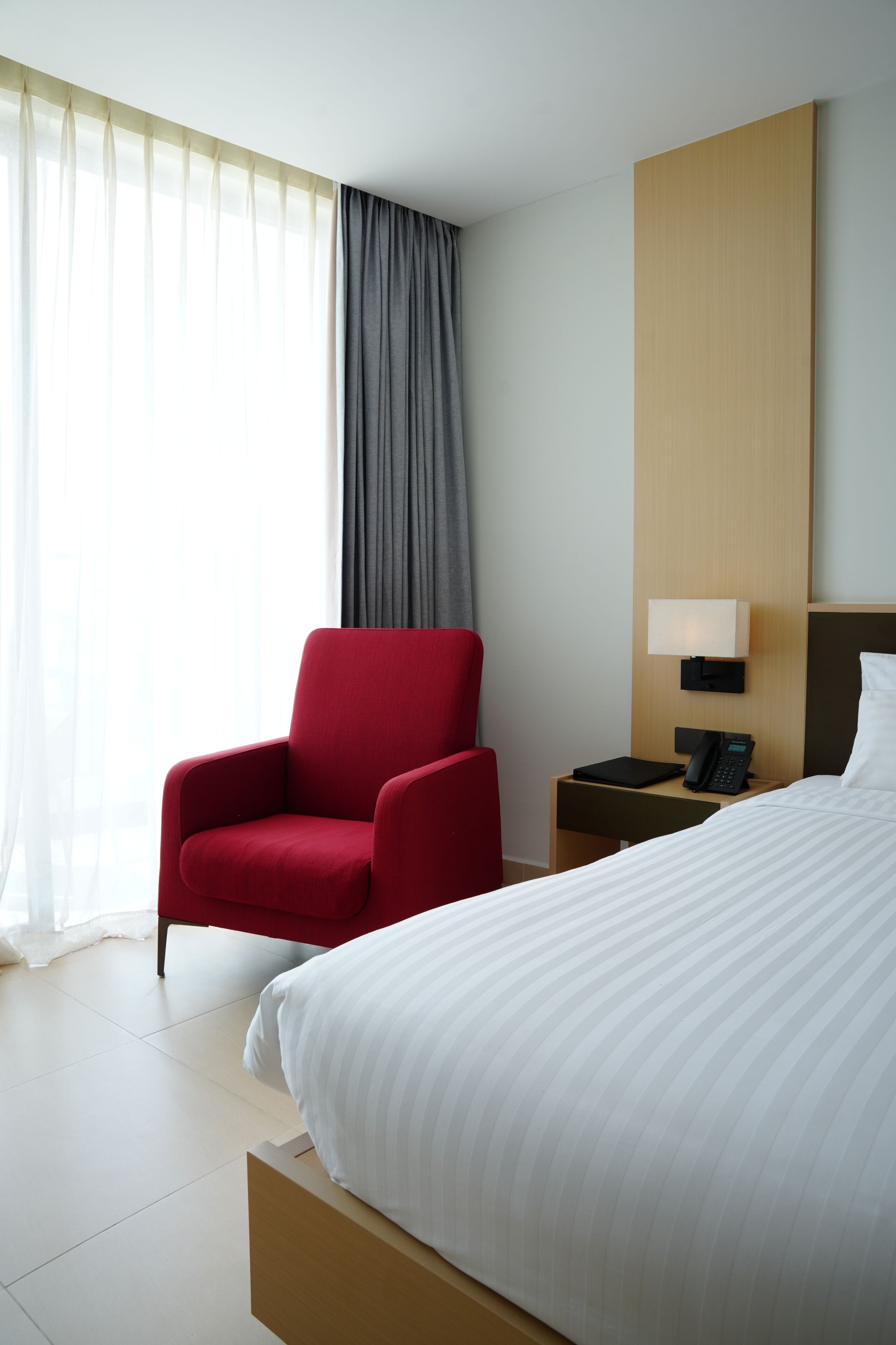 A hotel room with a bed, a red armchair, a bedside table with a lamp, and a window with curtains.