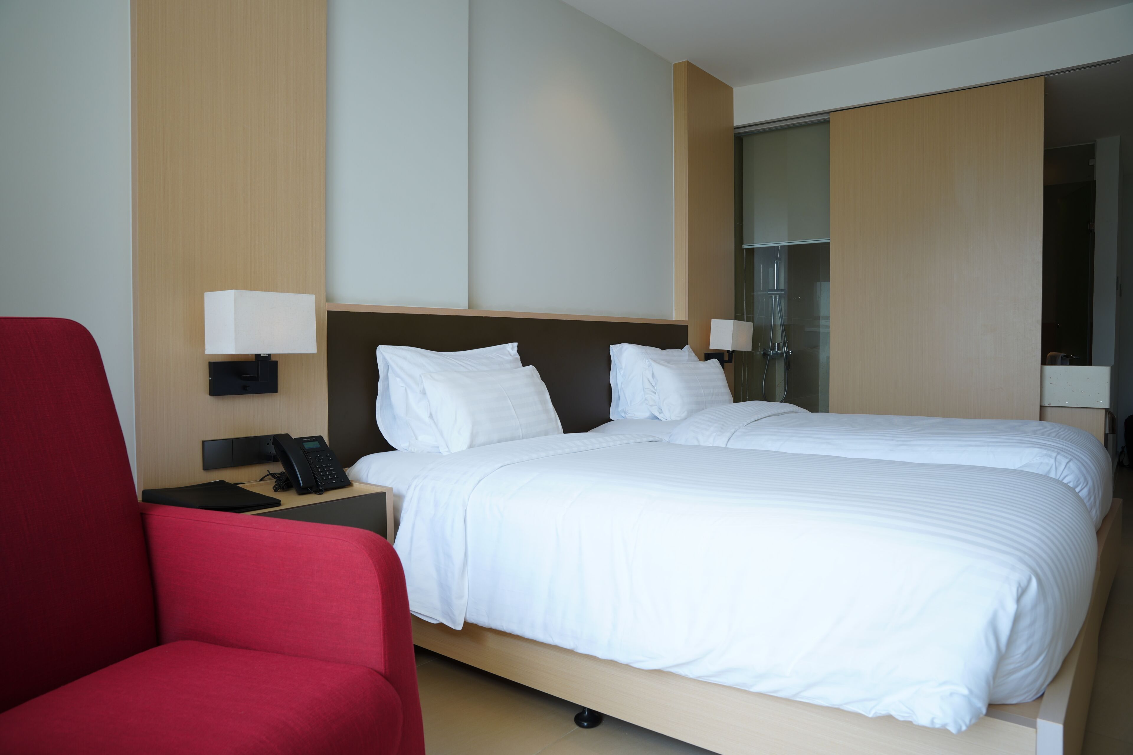 A hotel room with a bed, a desk with a phone, a red chair, and a nightstand with a lamp.
