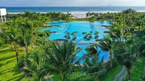 A large, circular swimming pool surrounded by palm trees, with a view of the ocean in the background. - Condotel Cam Ranh Beach Resort (Bac Cam Ranh)
