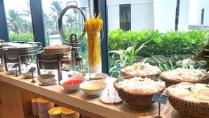 A buffet setup with various food items and utensils, including pasta, rice, and bowls, with a view of greenery and a building in the background. - Condotel Cam Ranh Beach Resort (Bac Cam Ranh)